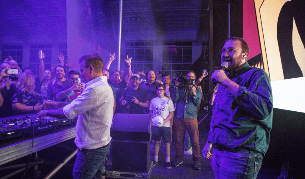 A man speaks into a microphone on stage beside a DJ at a blockchain event, while a lively crowd cheers, takes photos, and films the moment in a colorful, indoor concert setting.