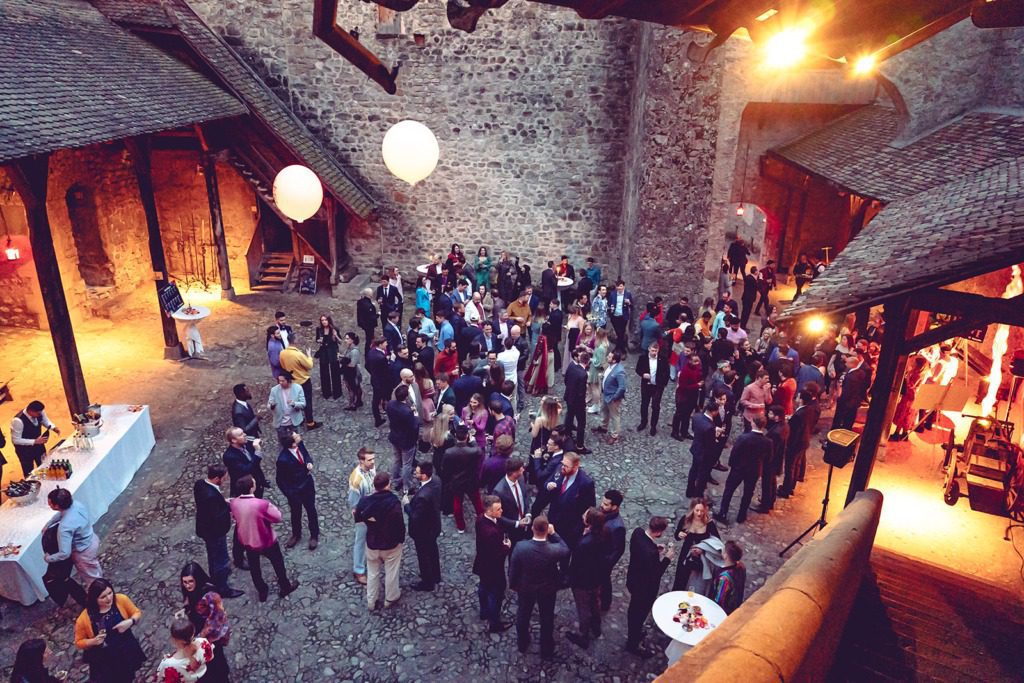 A large group gathers at an evening incentive travel event in a courtyard with stone walls. There are balloons, tables with drinks and food, and warm lighting, creating a festive atmosphere.