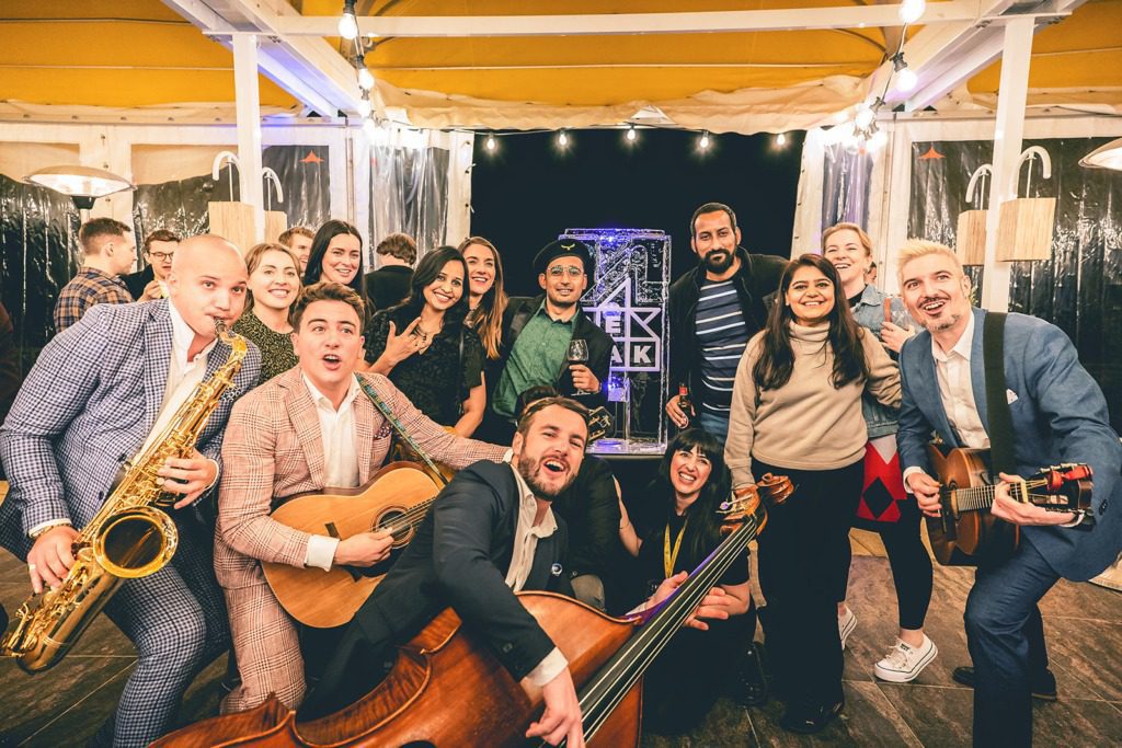 A group of people smiling and posing together indoors at an incentive travel event. Some are holding musical instruments including a saxophone, guitars, and a double bass. String lights and event decorations are visible in the background.