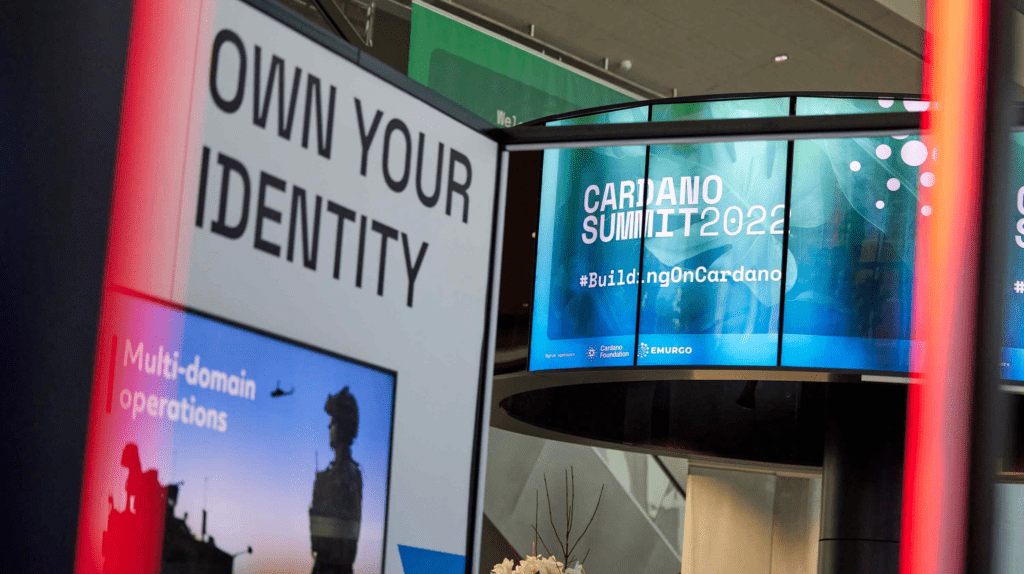 Photo of a digital display at a community event showing CARDANO SUMMIT 2022 #BuildingOnCardano, with a nearby sign reading OWN YOUR IDENTITY. A smaller screen highlights the text Multi-domain operations.