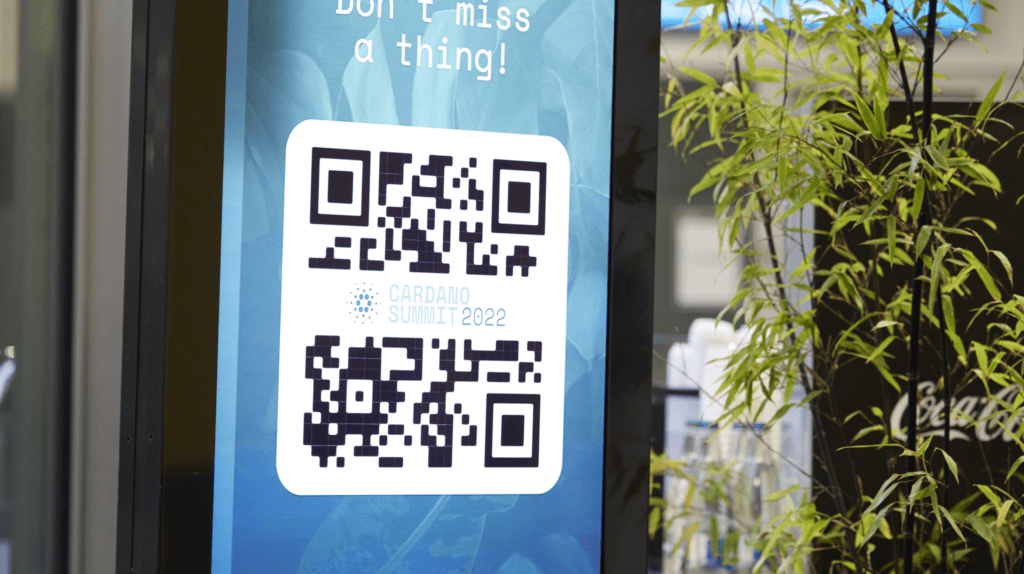 A digital sign displays a QR code with the text Don’t miss a thing! and CARDIAD SUMMIT 2022, highlighting this community event against a blue leafy background, next to a plant and a blurred Coca-Cola logo.