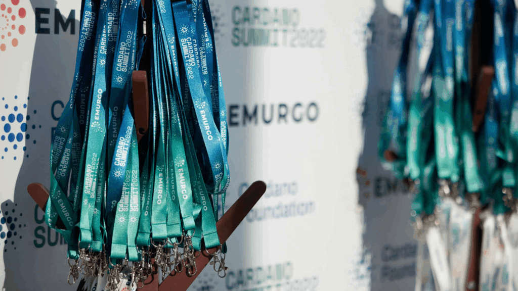 Close-up of teal and blue event lanyards hanging on a stand, with a blurred backdrop displaying Cardano Summit 2022 and EMURGO logos—capturing the atmosphere of a vibrant community event.