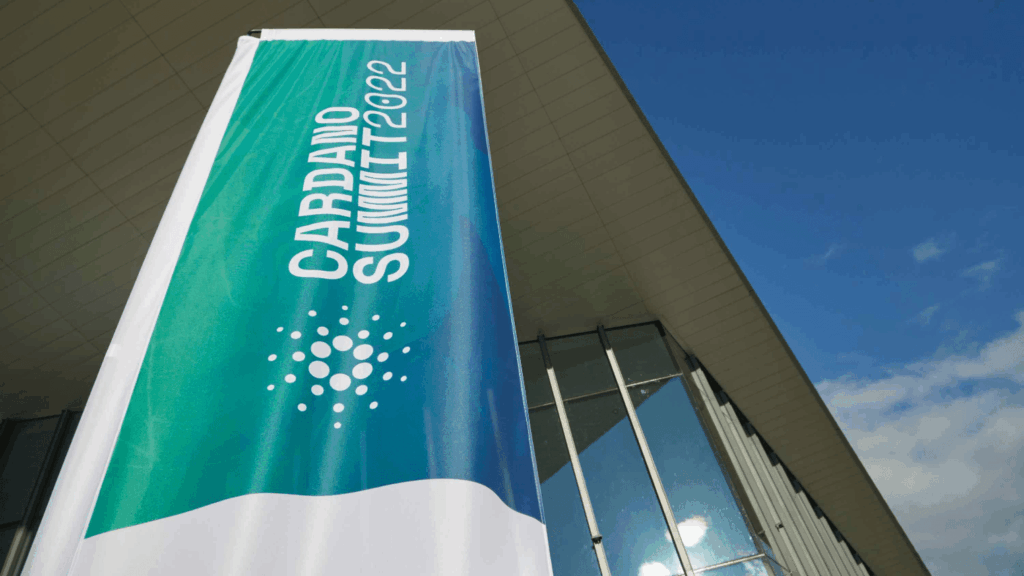 A vertical banner reading Cardano Summit 2022 with the Cardano logo stands in front of a modern glass building under a blue sky, highlighting the significance of this community event.
