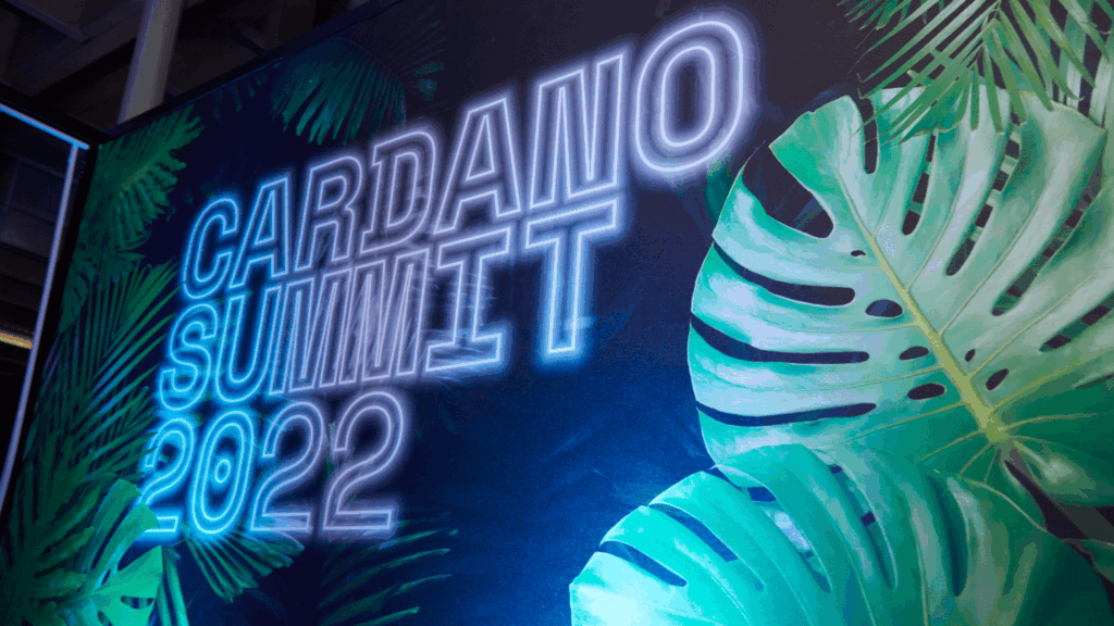 Neon-style text reads Cardano Summit 2022 against a dark background with large green tropical leaves, highlighting this vibrant community event.