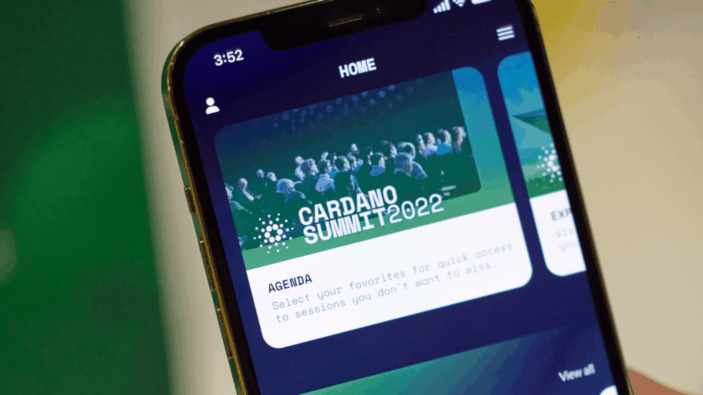 A smartphone screen displays an app page for Cardano Summit 2022, highlighting a vibrant community event with a group of people on stage and event details in a green-themed design.
