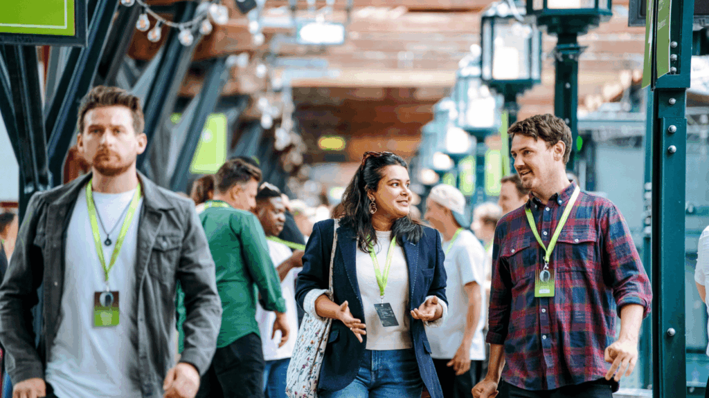 A group of people walk through a busy sustainability & diversity conference. Two attendees wearing green lanyards chat and smile, while others walk in the background. The atmosphere appears social and lively.