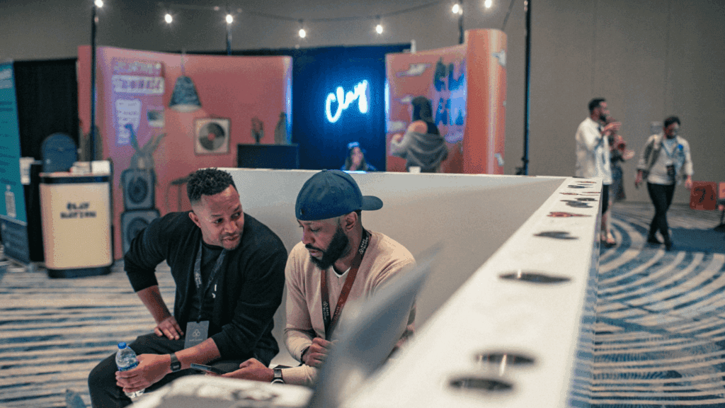 Two men sit and talk at an experiential marketing event booth with colorful displays and a neon “Chuy” sign in the background, while others mingle in the lively event space.