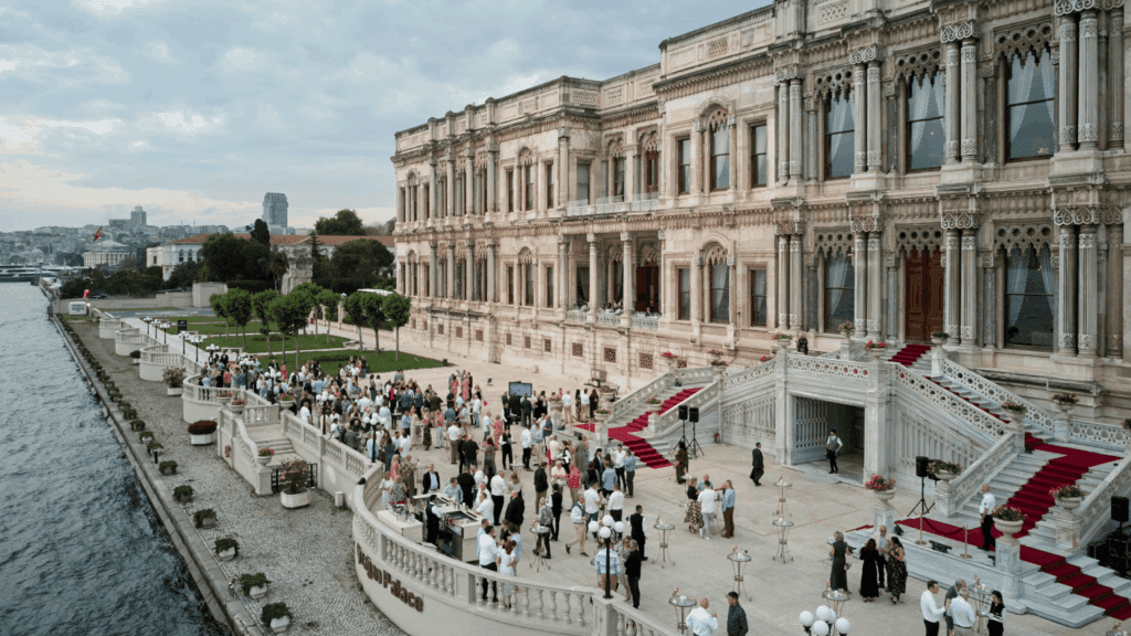 A large crowd gathers for a conference on the elegant marble terrace of a grand, historic waterfront palace with intricate architecture, red carpets on the stairs, and a scenic river beside the building under a cloudy sky.