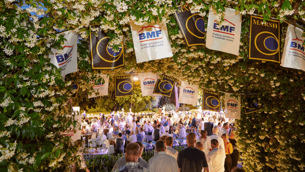 A large outdoor conference unfolds under an archway adorned with flowers and banners, with many people seated at tables and mingling in a festive, garden-like setting.