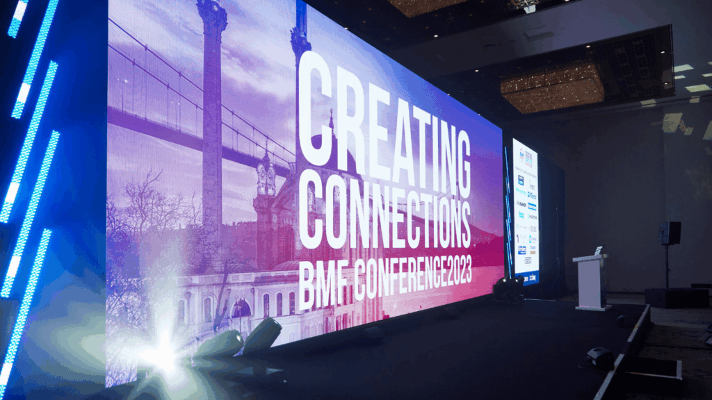 A large digital display at the conference reads CREATING CONNECTIONS BMF CONFERENCE 2023 in a spacious, modern event hall with a podium and screens.