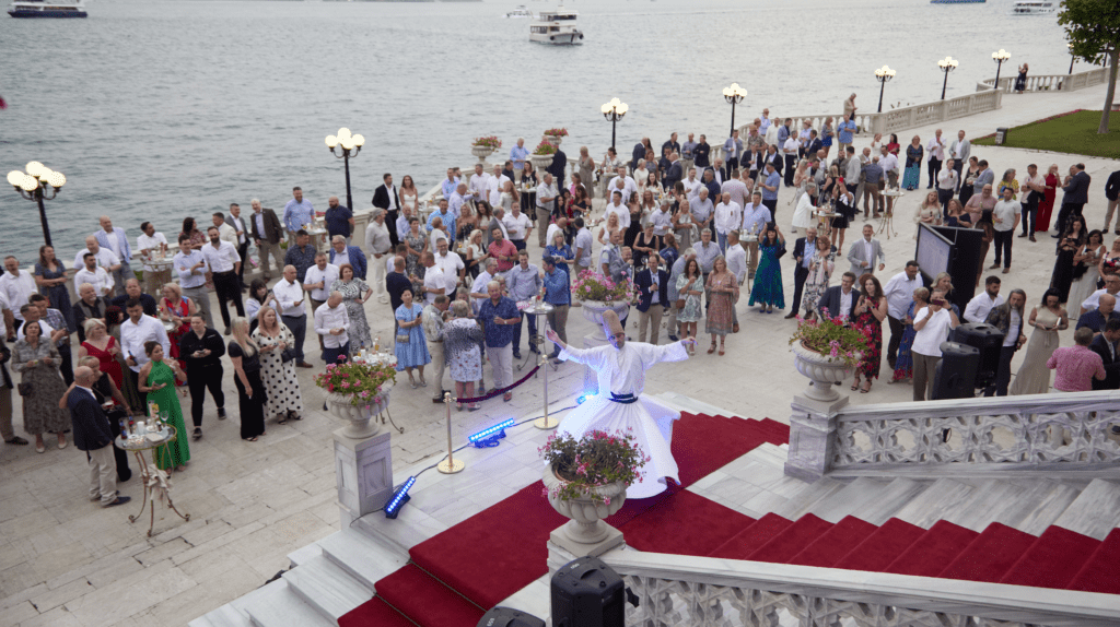 A large group gathers at an outdoor waterfront conference venue, watching a whirling dervish perform on red-carpeted stairs. Boats glide by in the background, while ornate stone railings and potted flowers enhance the vibrant scene.