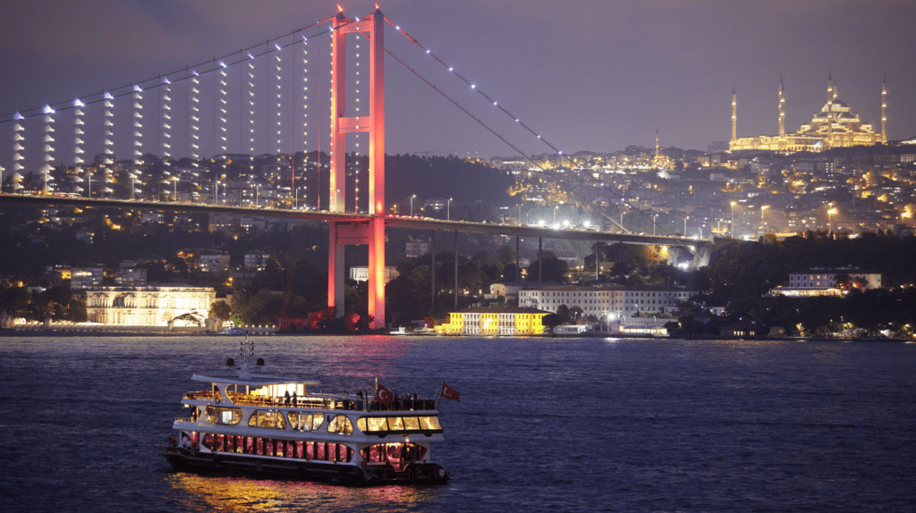 A brightly lit conference boat sails on the Bosphorus at night in Istanbul, with the illuminated Bosphorus Bridge and a mosque on a hill in the background. City lights and buildings are visible along the waterfront.