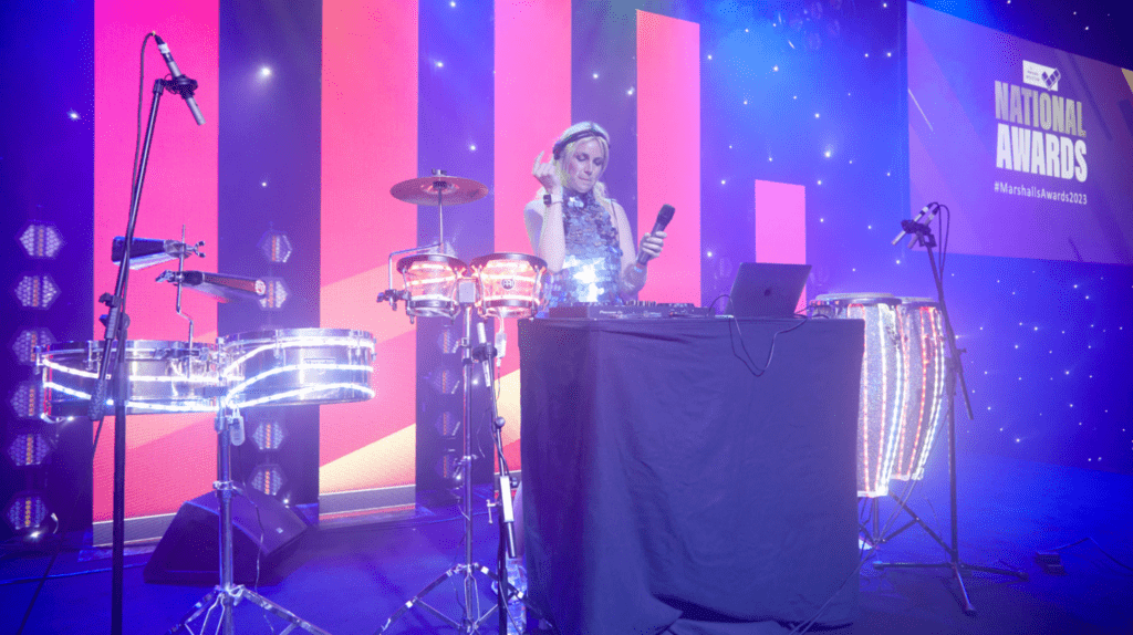 A DJ stands behind a table with a laptop and mixer, playing music on stage with electronic drums and congas, surrounded by colourful lights during a Roadshow, with a screen displaying National Awards in the background.
