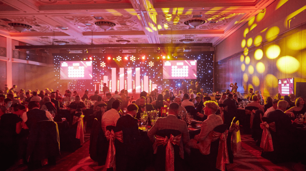 A large, elegant banqueting hall filled with people seated at round tables, decorated with gold chair sashes. Bright stage lights and illuminated screens create a festive atmosphere at an evening roadshow or awards ceremony.