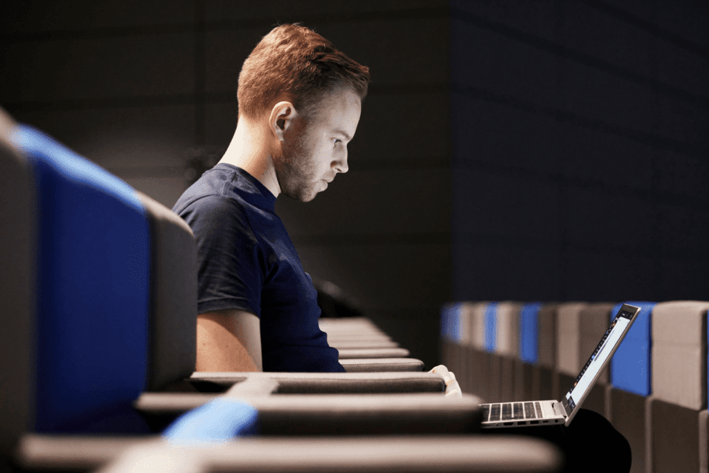 A man sits alone in a row of modern auditorium seats, focused intently on his open laptop, illuminated by the screen’s light as he explores AI in events in an otherwise dimly lit room.