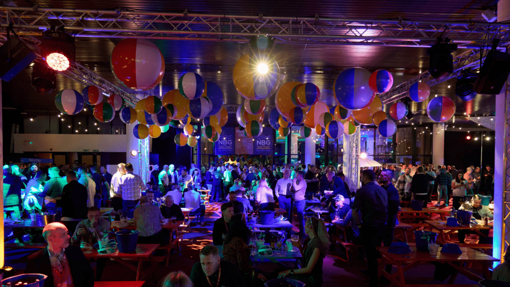 A large, colorful indoor conference & exhibition with many people socializing at long red tables, under hanging beach balls and festive lighting, creates a lively party atmosphere.