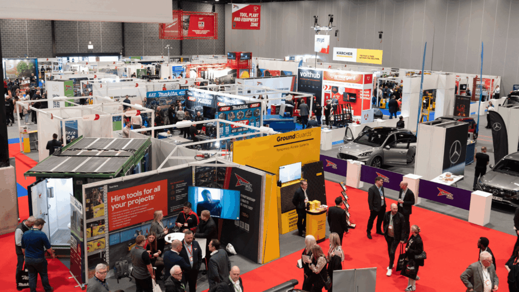 A busy Speedy Hire Live Expo with many booths, displays, and attendees walking around. The scene features cars, banners, digital screens, and product stands in a large indoor exhibition hall with red carpeted walkways.