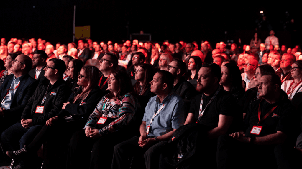 A large audience, many wearing name badges, sits closely together in a dimly lit venue at the Speedy Hire Live Expo, attentively watching a presentation as reddish lighting casts a warm hue across the crowd.