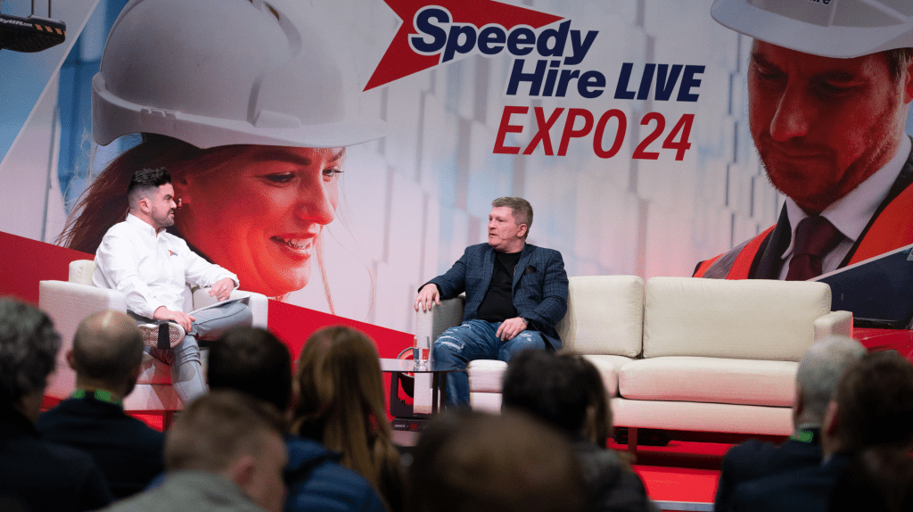 Two men sit and talk on stage at the Speedy Hire Live Expo 24 event. One wears a white shirt, the other a checked jacket. The stage backdrop features people in safety helmets and the Live Expo logo as audience members face the stage.