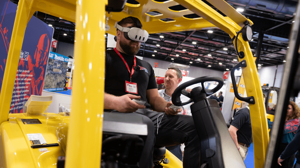 At Live Expo, a man sits in the driver’s seat of a yellow construction vehicle, wearing a VR headset and holding controllers, while another man stands nearby in a busy indoor exhibition setting.