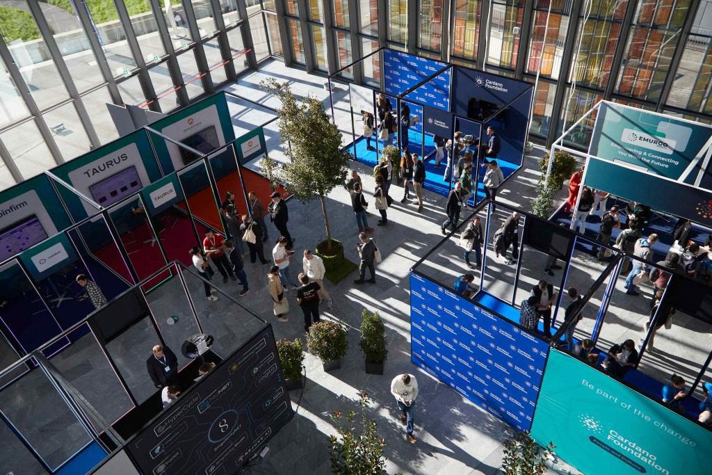 Aerial view of a modern conference & exhibition featuring booths, banners, and groups of people networking in a spacious, sunlit hall with large windows and potted trees.