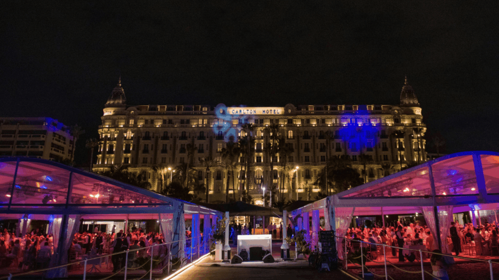 A grand, illuminated hotel at night is seen in the background, while in the foreground, people gather and dine under lit tents, celebrating a sales incentive with a festive atmosphere and colorful lighting.