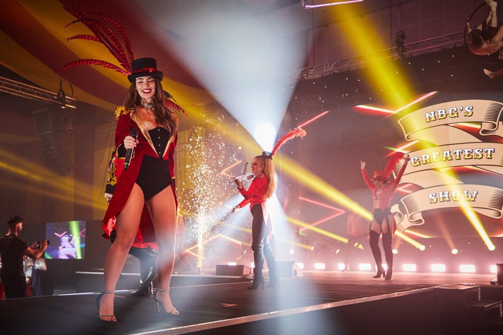 A smiling female performer in a red circus ringmaster costume stands on a stage with bright lights and confetti. Two other performers dance behind her, as “NBC’s Greatest Show” shines in the background—an experience crafted by a creative event agency.