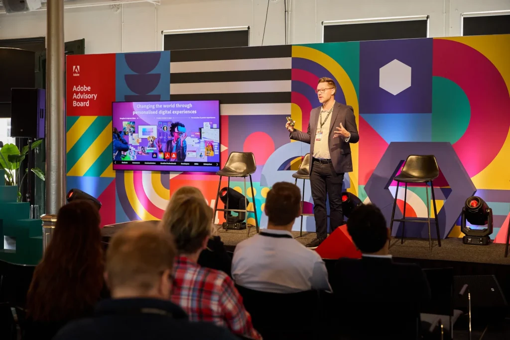 A man in business attire speaks on stage at an Adobe Advisory Board event, presenting creative content beside a screen and a colorful geometric backdrop, while an audience listens attentively in the foreground.
