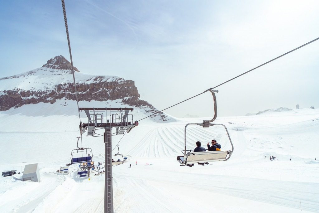 People enjoy ski lift experiences over a snowy mountain landscape, with others skiing and snowboarding below; a tall, rocky peak rises in the background under a bright, cloudy sky.