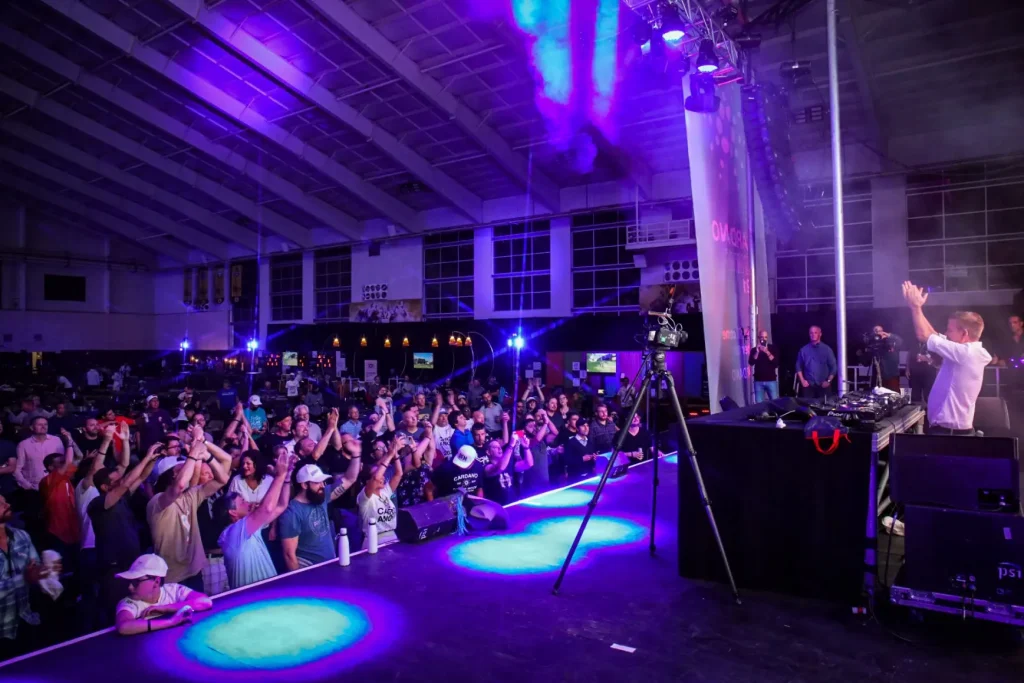 A DJ performs on stage in a large indoor venue with purple lighting, as a crowd cheers and takes photos. A camera on a tripod records the event—an energetic scene that showcases effective event strategy in action.