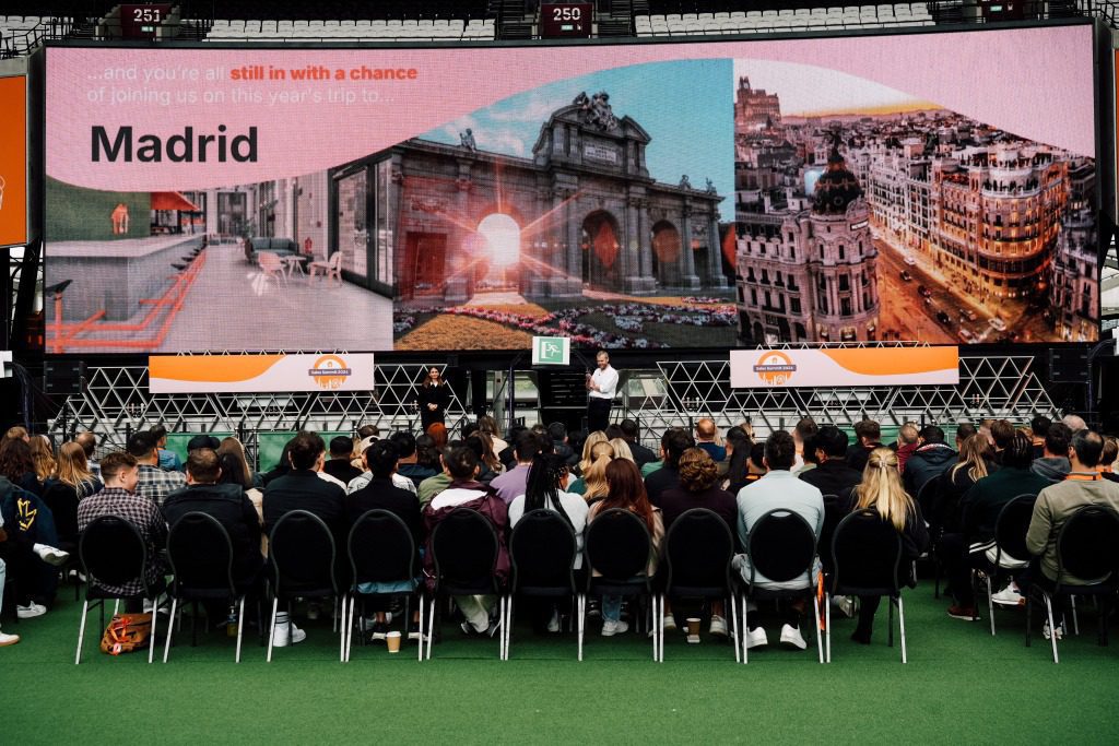 A crowd sits facing a stage where two speakers stand, with a large screen behind them displaying “Madrid” and city landmarks—an inspiring outdoor event on green turf, bringing strategy to event marketing in a vibrant setting.