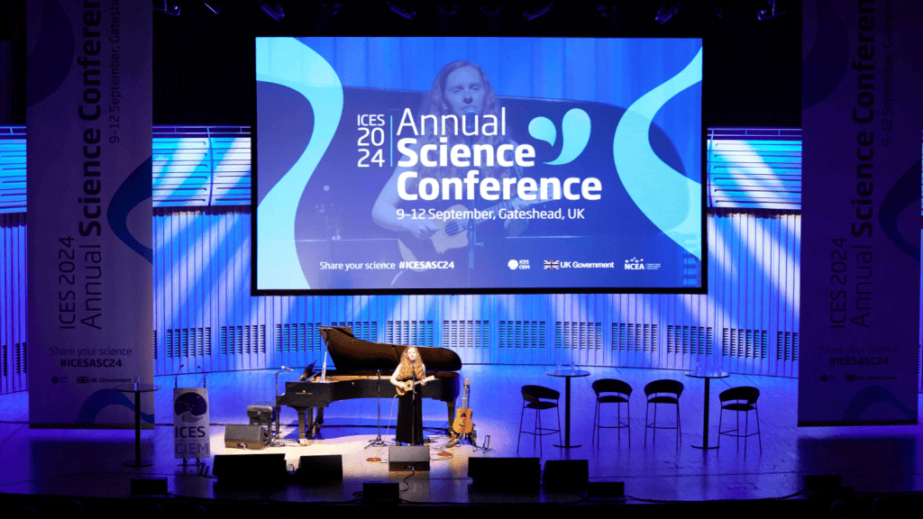 A stage set for the science Conference features a grand piano, microphones, guitar, and empty chairs, with a large conference banner and event details displayed on a screen above.