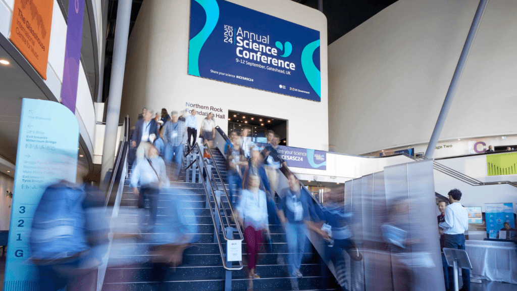 A group of people moves up and down a busy staircase inside a modern building, with a large sign above reading Science Conference and details about the event. The scene is energetic and bustling.