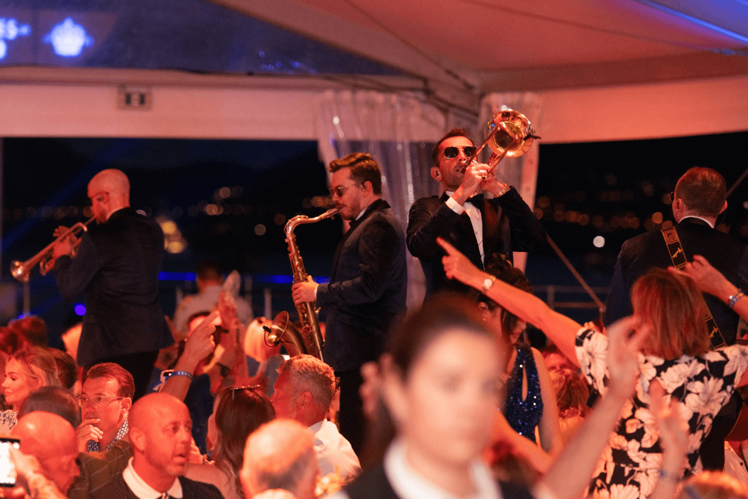 A jazz band performs on stage at a lively indoor party, meeting events demand as guests dance and raise their hands. The musicians play trumpet, saxophone, and trombone under warm, colorful lighting.