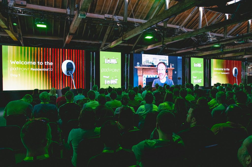 A large audience watches a conference presentation in a dimly lit venue with wooden beams. Multiple screens showcase Tech at Events, displaying a welcome message for podPOINT Resident 2023 and a video call with a smiling speaker.