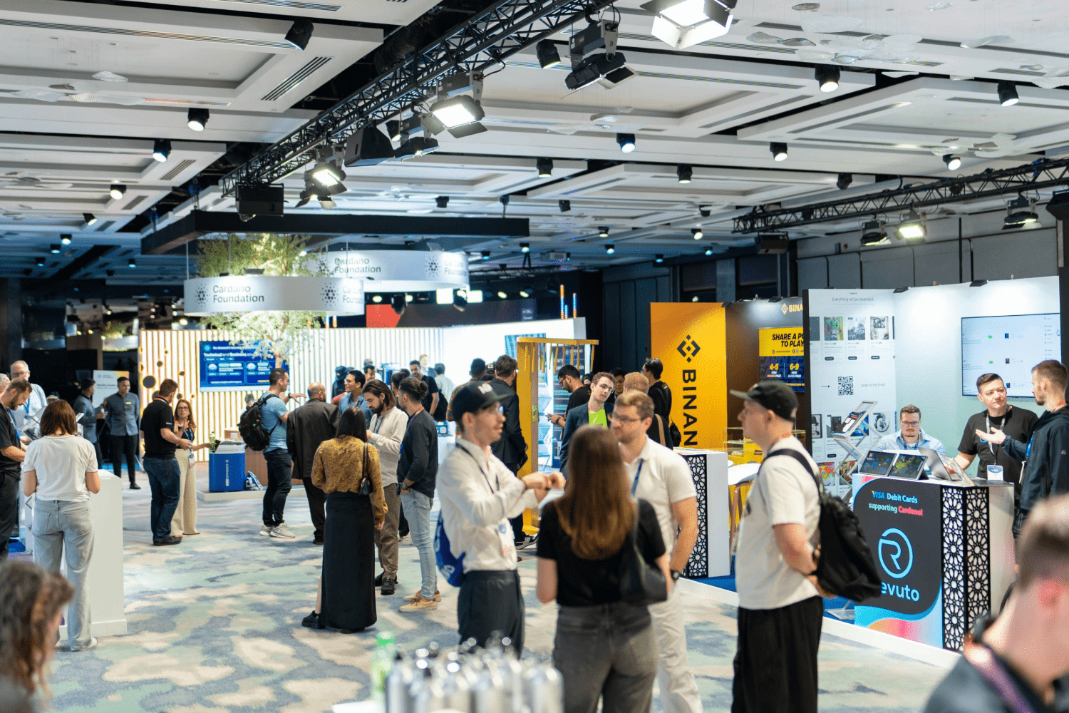 People gather and network at a busy tech conference or expo, enjoying live experiences among various exhibition booths, including one for Binance, in a large, well-lit event hall.