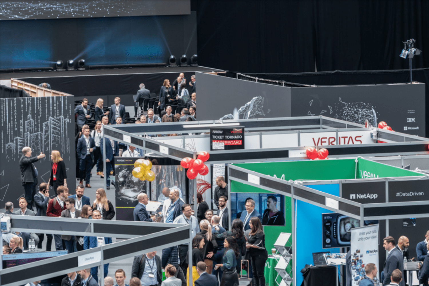 A busy tech conference with people walking and talking among several exhibition booths decorated with signs, banners, and balloons, inside a large indoor venue.