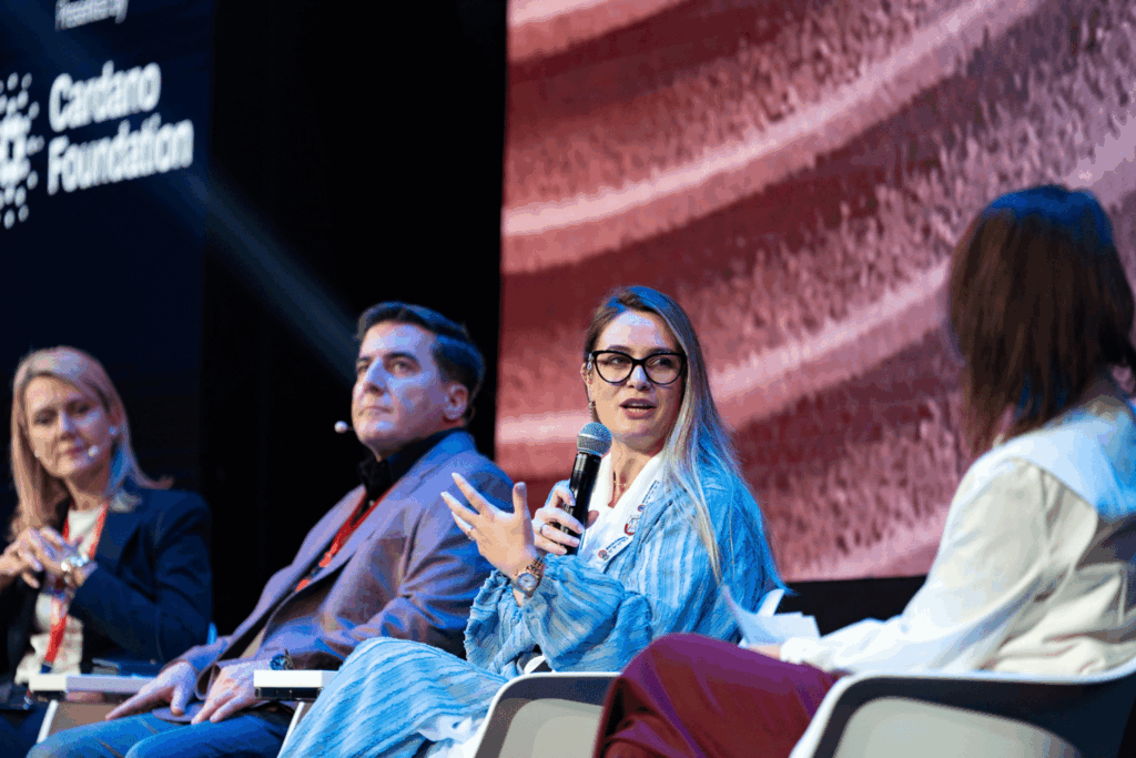 Four people sit on a panel discussion stage. A woman in the foreground speaks while gesturing with her hand. The panelists, involved in Event Marketing & Communications, have name badges and a “Cuatro Foundation” logo is visible in the background.