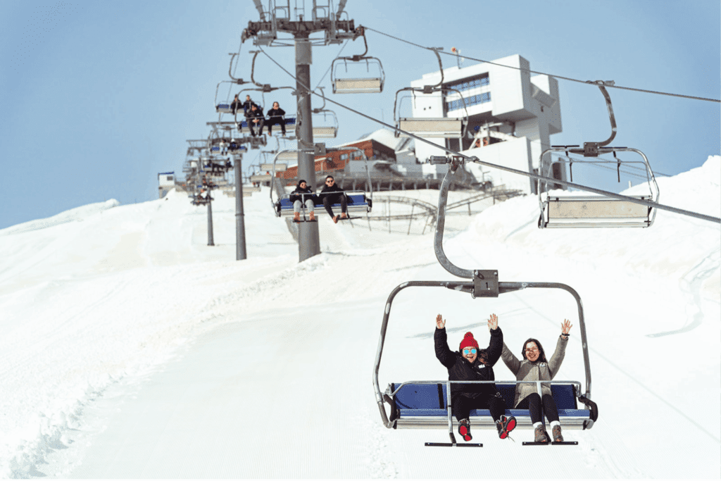 People ride a ski lift up a snowy mountain on a sunny day, enjoying the excitement of incentive events. A few buildings are visible at the top under a clear blue sky, while the front two riders smile and wave.