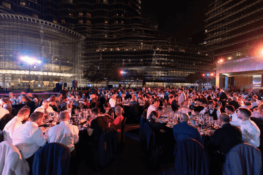 A large crowd of people seated at round tables enjoys a formal outdoor dinner event at night, surrounded by modern city buildings with bright lights—an impressive display of Event Marketing Services in action.