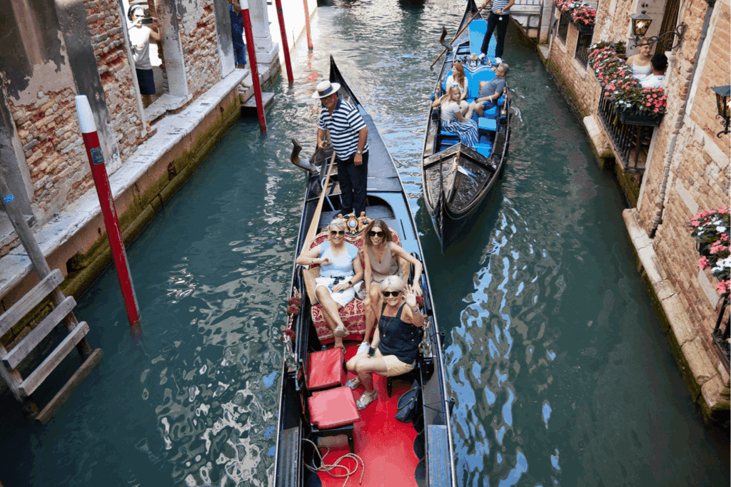 A group of people enjoy a gondola ride through a narrow Venice canal during exciting Incentive Events, passing brick buildings with flowers, while another gondola with passengers follows behind.