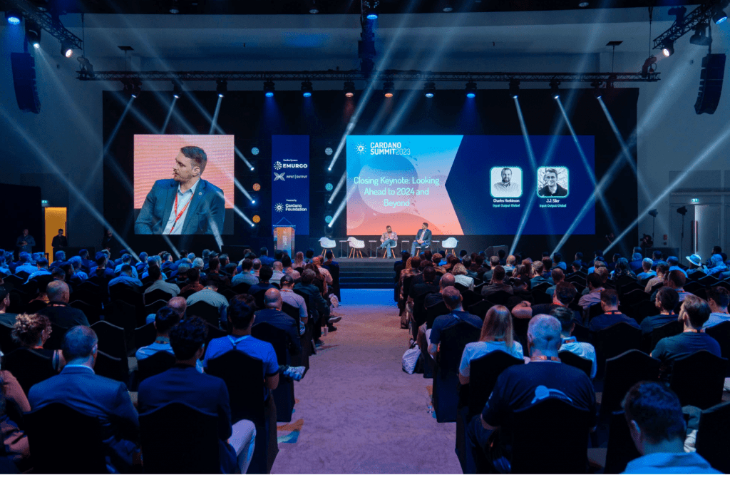 A large audience sits in a conference hall facing a stage with two speakers in discussion. The stage, typical of top conference venues, is brightly lit with spotlights and features large screens displaying event branding and speaker information.
