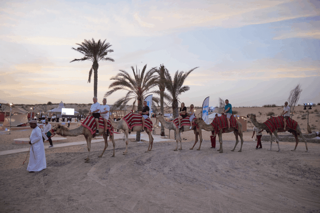 A group of people ride camels draped in red and white blankets along a sandy path lined with palm trees at sunset during exclusive incentive events, while guides in traditional clothing stand nearby.