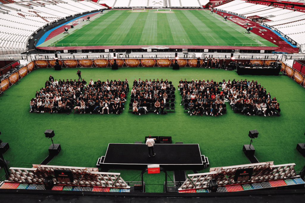 A speaker addresses a large audience seated on green turf in a stadium, gathered for an Event Marketing & Communications seminar. The audience is arranged in three distinct rectangular groups, with empty stands and a football field visible in the background.