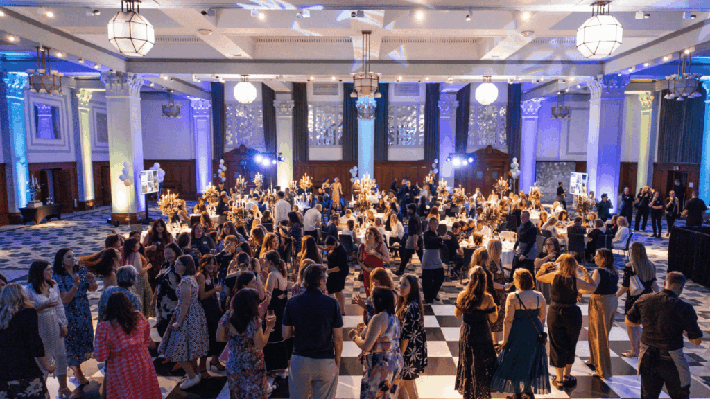 A large crowd of people dressed in formal attire socializes and dances at a private luxury event in an elegant, spacious ballroom decorated with chandeliers, blue lighting, and checkered flooring.