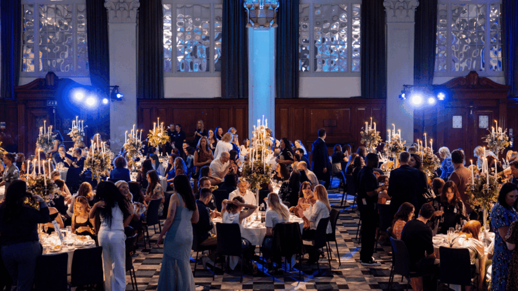 A large, elegant banquet hall hosts a private luxury event, with well-dressed guests seated at round tables adorned with tall, flower-filled candelabras. Blue lighting and ornate windows enhance the formal, festive atmosphere.