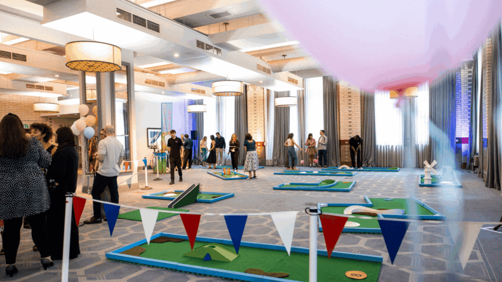 People play mini-golf on colorful courses set up inside a large, bright room decorated with balloons and bunting, perfect for a private luxury event. Groups mingle and watch, creating a lively, festive atmosphere.