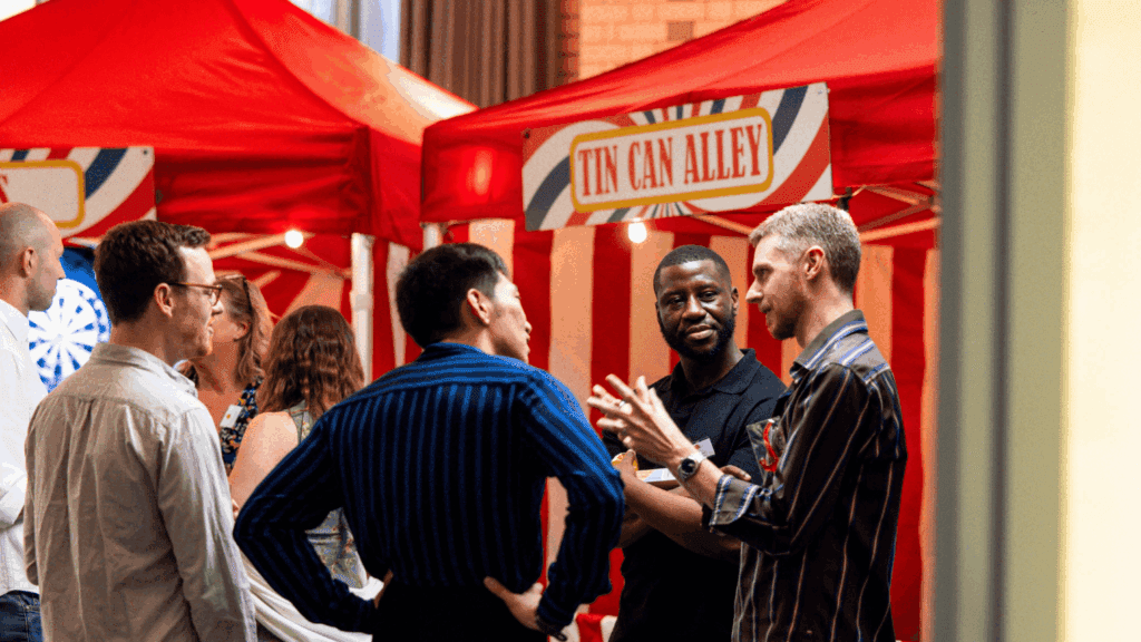 A group of people stand and chat in front of red and white carnival tents at a private luxury event. A sign reading Tin Can Alley hangs above one tent. The atmosphere appears lively and casual.