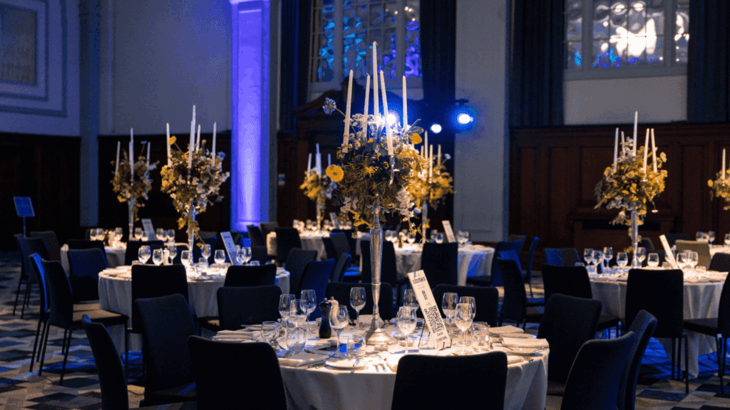 A formal dining hall set for a private luxury event, with round tables draped in white cloths, elegant glassware, tall floral centerpieces with candles, and blue accent lighting creating a sophisticated atmosphere.