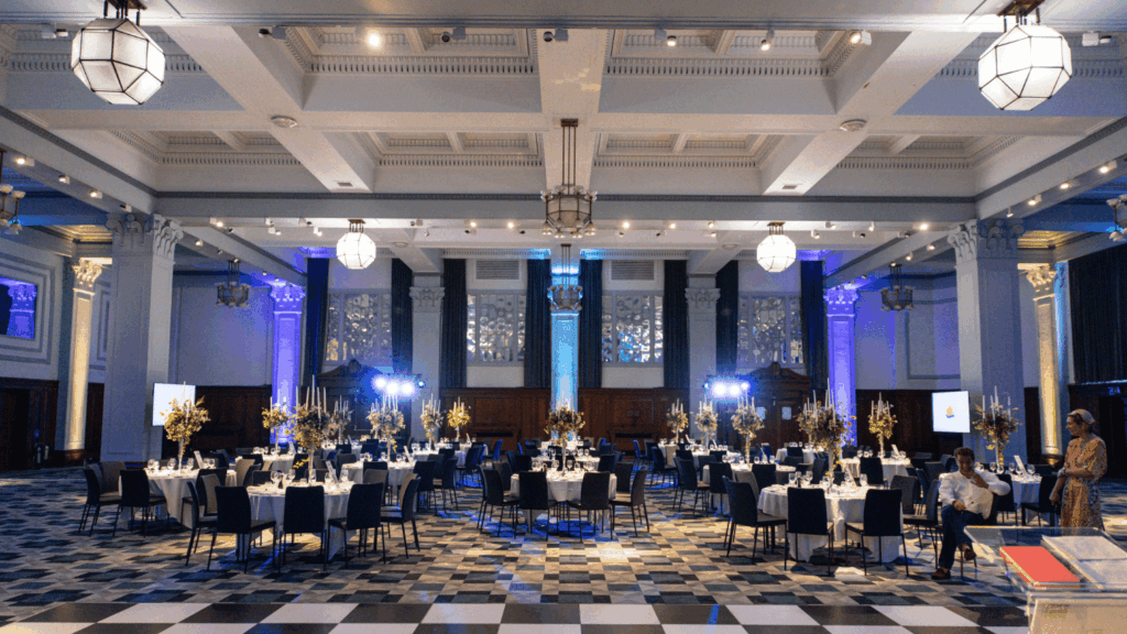 A large, elegant banquet hall with high ceilings, chandeliers, and floral centerpieces is set for a private luxury event. Round tables and a checkerboard floor shine under blue lighting as a few people prepare the sophisticated space.